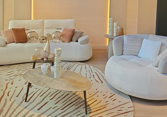 Ambiance cocooning avec canapé blanc aux courbes douces, fauteuil arrondi et table basse ovale en pierre, sur un tapis rond aux motifs beiges et rosés