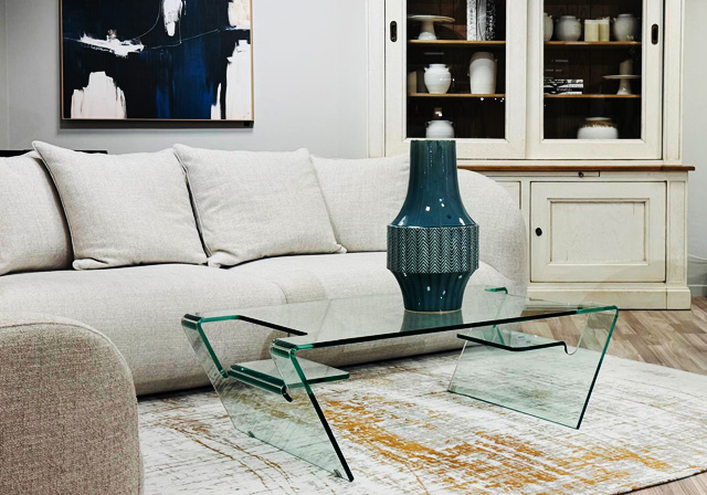 Salon élégant au magasin Mobilier de France Saint-Lô, avec canapé beige clair, table basse design en verre transparent et grand vase bleu canard, sur fond de meuble vitrine blanc en bois patiné
